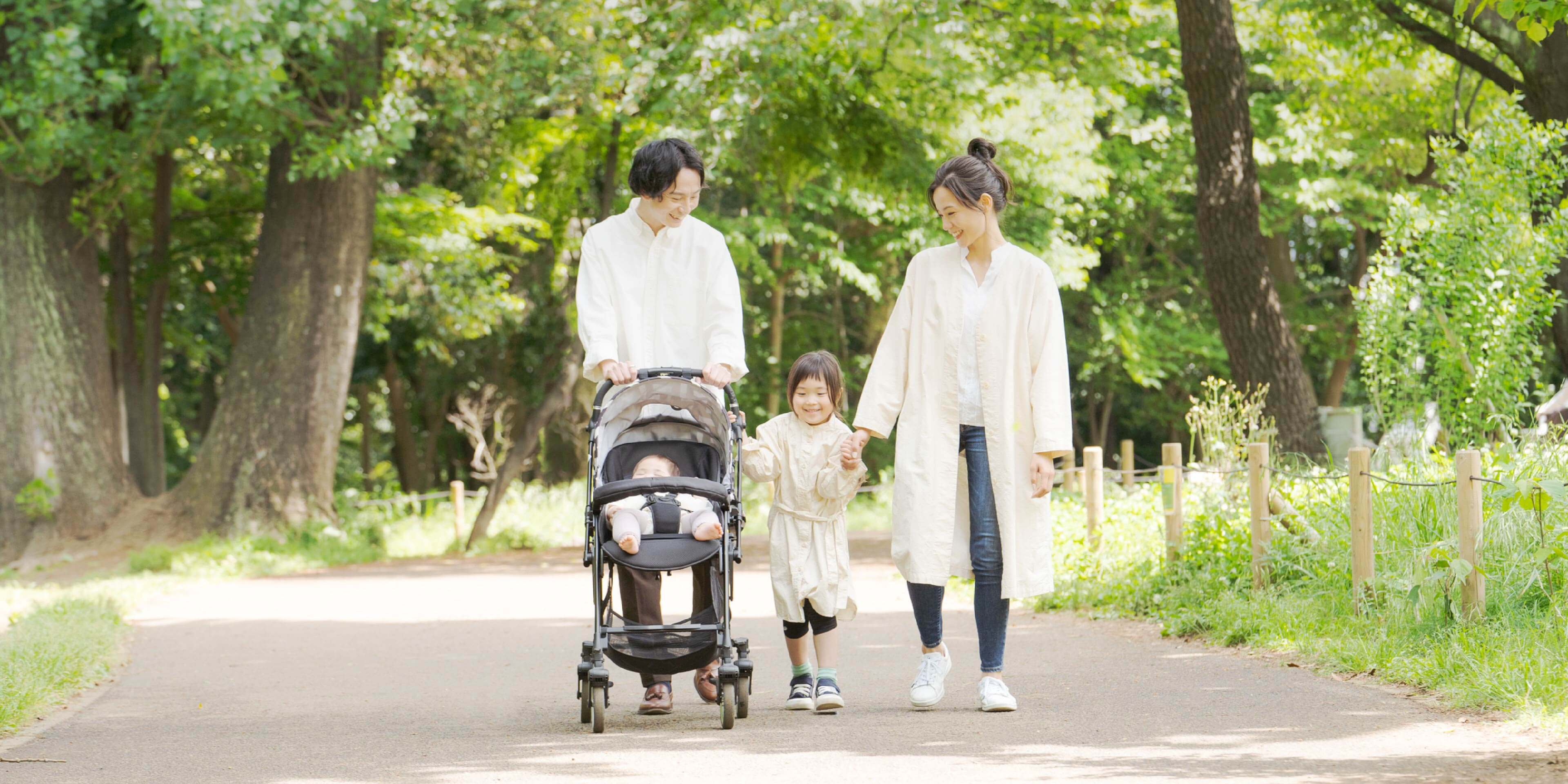 家族で散歩している。父親がベビーカーを押し母親と子どもが手を繋いでいる。